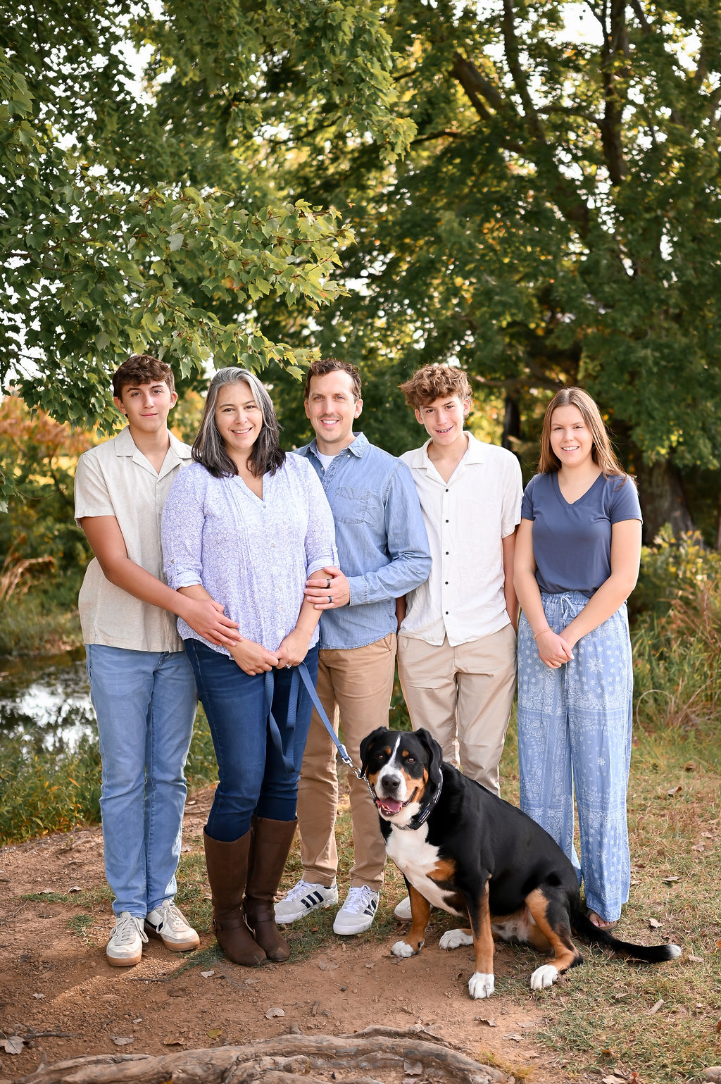Sarah posing outdoors with her husband, three children, and their large black-and-tan dog. Sarah is wearing a light purple patterned blouse and dark jeans, standing under a large leafy tree with her family in a sunny, natural landscape. 