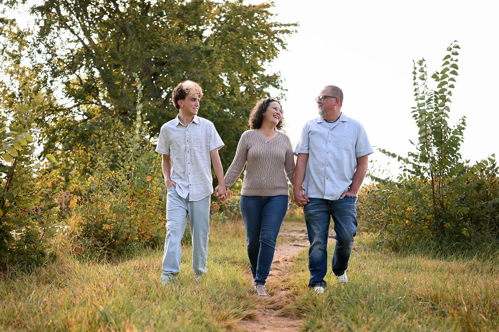 Heather walking along a grassy trail with her husband, Brandon, and son, Aidan. They are holding hands and smiling at one another, set against a backdrop of golden-hour sunlight and lush trees. 