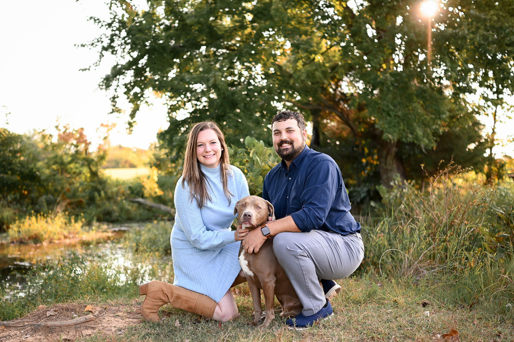A man with short dark curly hair and a beard smiles while crouching outdoors beside a woman with long brown hair and a light blue turtleneck sweater dress. They are posing together with a brown mixed-breed dog in a sunlit outdoor setting with trees, tall grass, and a small pond in the background. 